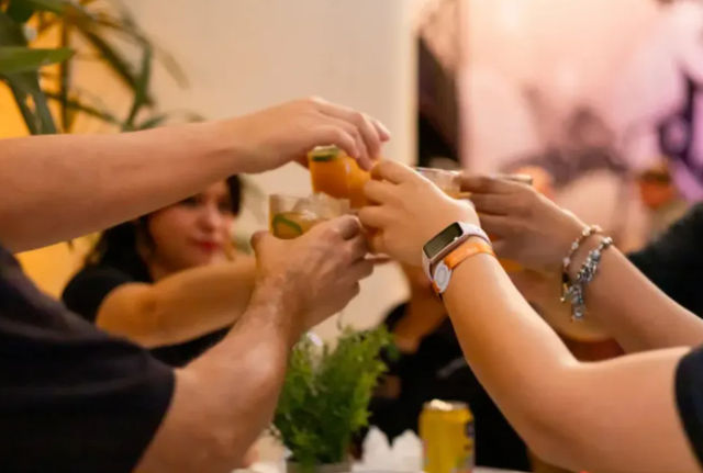 Group of people clinking orange citrus cocktails in a lively indoor toast, close-up of hands and drinks with blurred guests and potted plant in background.