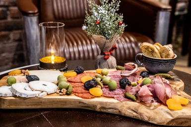 Candlelit charcuterie spread on a rustic wooden board: sliced cheeses, salami and prosciutto, green olives, cornichons, dried apricots, nuts and toasted baguette slices on a cozy lounge table with a small festive potted plant.
