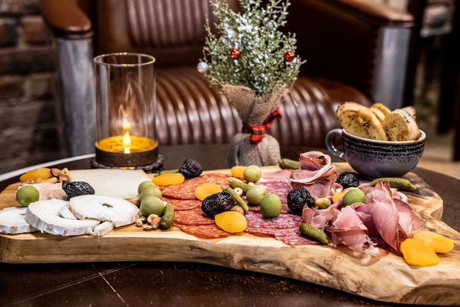 Candlelit charcuterie spread on a rustic wooden board: sliced cheeses, salami and prosciutto, green olives, cornichons, dried apricots, nuts and toasted baguette slices on a cozy lounge table with a small festive potted plant.