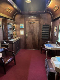 Cozy vintage wood-paneled sleeper cabin interior in a train carriage with red carpet, leather chairs, luggage trunk and display shelves.