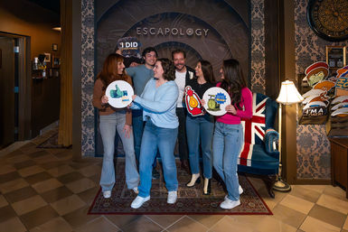 Six people celebrating in an escape room lobby, posing with themed achievement signs and props in front of a decorative patterned backdrop and a Union Jack armchair.