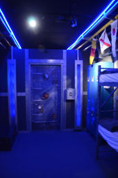 Blue-lit nautical-themed interior featuring a riveted steel submarine hatch door, metal lockers and bunk beds, LED strip lighting and hanging signal flags.