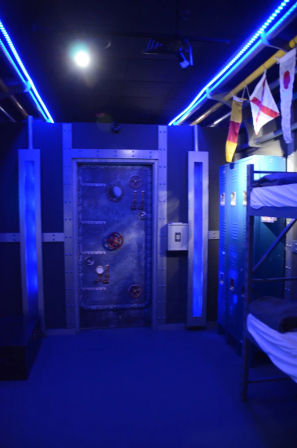 Blue-lit nautical-themed interior featuring a riveted steel submarine hatch door, metal lockers and bunk beds, LED strip lighting and hanging signal flags.