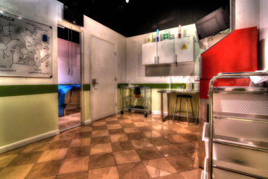 Bright clinical laboratory prep room with checkered tile floor, white cabinets bearing biohazard symbols, stainless rolling cart, stool and a chemistry whiteboard