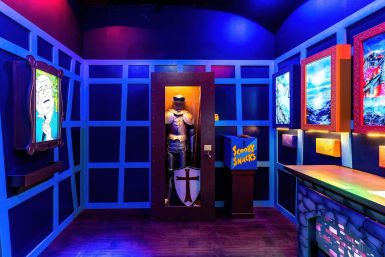 Neon-lit, Scooby-Doo–themed attraction interior featuring a knight in a glass case, a blue 'Scooby Snacks' box on a pedestal, colorful framed artwork, and purple-blue grid-patterned walls — playful family entertainment set design.