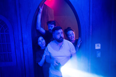 Four adults huddle in an arched wooden doorway of a blue-lit escape room or haunted attraction; a bearded man holds a bright flashlight beam through fog as the group peers ahead with tense, curious expressions.