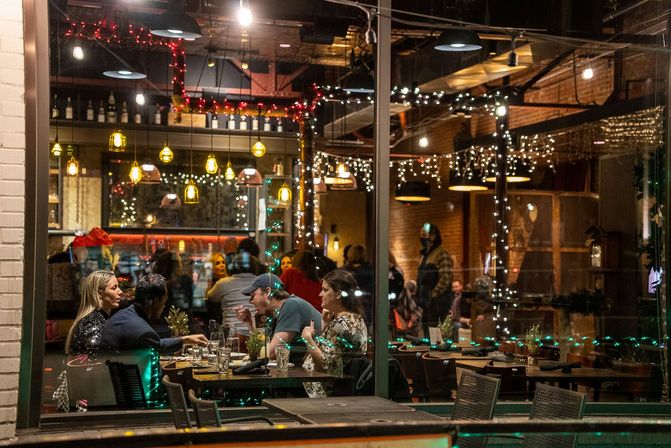 Cozy urban restaurant at night seen through a window — friends sharing dinner at a table under warm pendant and string lights, exposed brick interior and a lit bar in the background.
