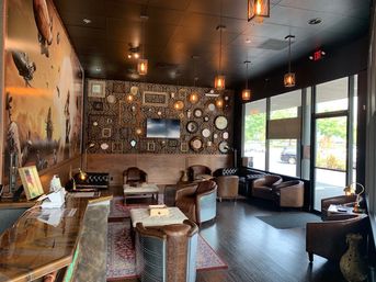 Cozy urban lounge interior with leather armchairs and tufted sofas, patterned accent wall of clocks and frames, hanging lantern lights and large storefront windows.