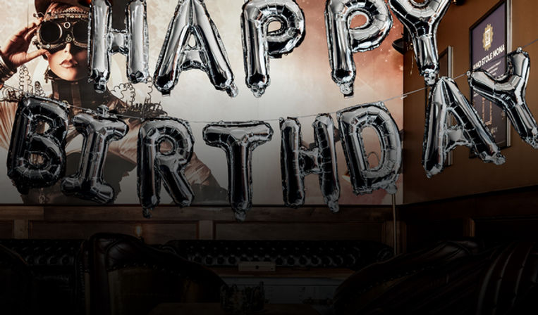 Silver foil 'HAPPY BIRTHDAY' letter balloons hanging across a dimly lit lounge with dark leather sofas and a steampunk-style mural on the wall.