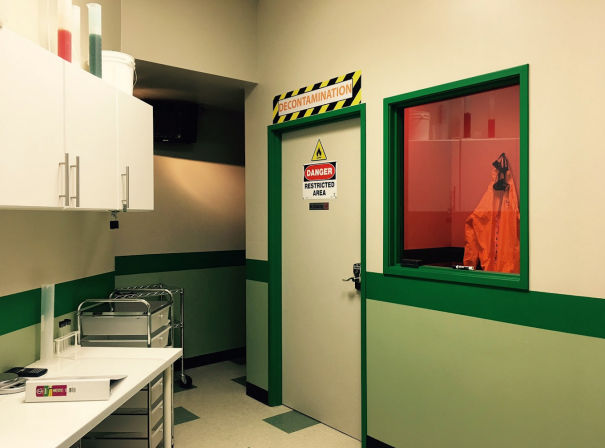 Indoor lab corridor with green-trimmed decontamination door labeled Danger Restricted Area, red-tinted window revealing an orange hazmat suit, white cabinets and stainless cart.