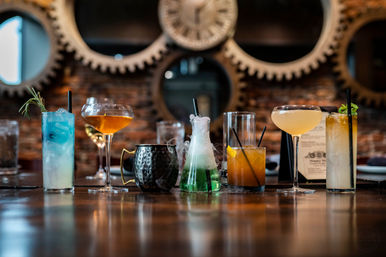 Colorful craft cocktails lined up on a polished wooden bar in an industrial-style setting with exposed brick and large gear wall décor — blue highball with rosemary, amber coupe glasses, a hammered mule mug, a smoky green beaker cocktail, an old-fashioned, and a mint-topped tall drink.