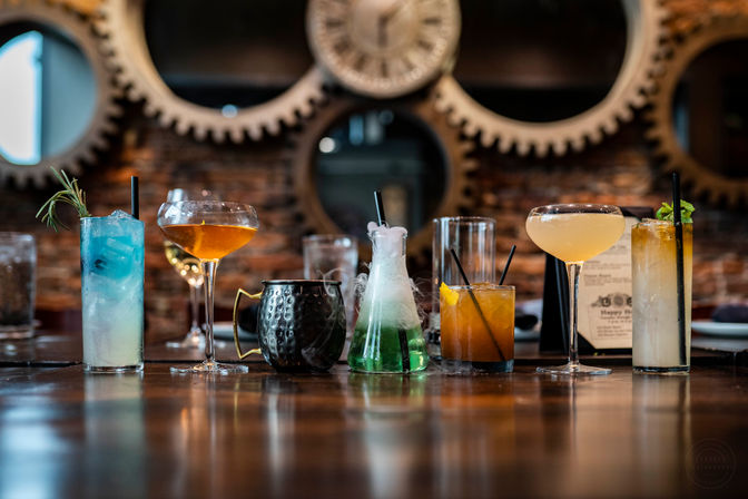 Colorful craft cocktails lined up on a polished wooden bar in an industrial-style setting with exposed brick and large gear wall décor — blue highball with rosemary, amber coupe glasses, a hammered mule mug, a smoky green beaker cocktail, an old-fashioned, and a mint-topped tall drink.
