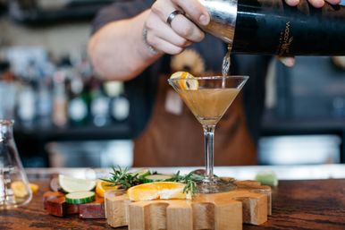 Bartender pouring a craft cocktail from a shaker into a martini glass on a wooden bar countertop, garnished with orange slice, lime and rosemary—artisan cocktail at a cozy bar.