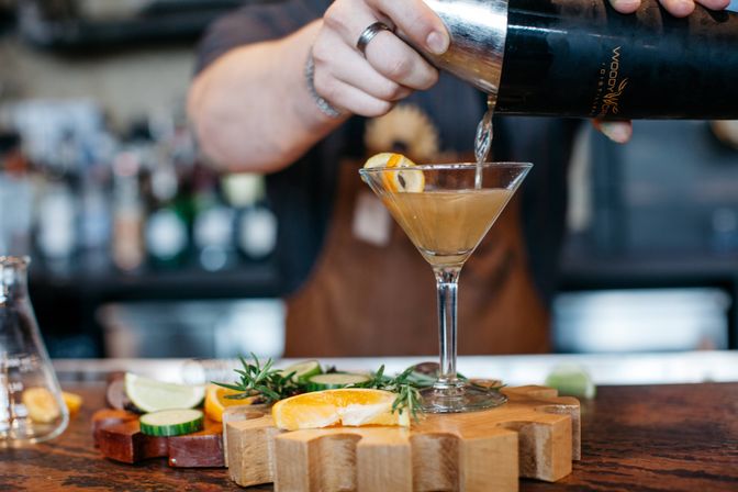 Bartender pouring a craft cocktail from a shaker into a martini glass on a wooden bar countertop, garnished with orange slice, lime and rosemary—artisan cocktail at a cozy bar.