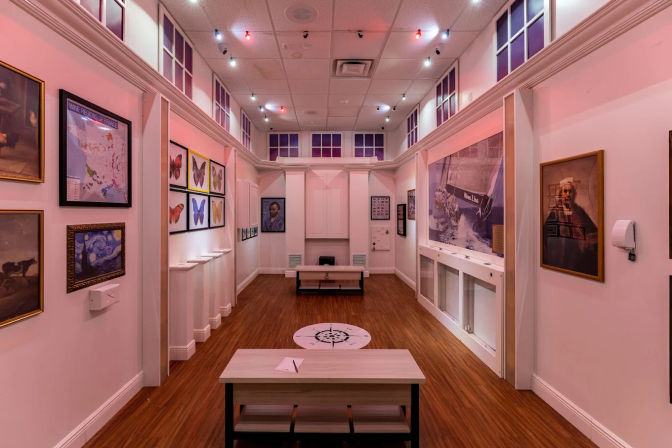 Narrow indoor art gallery with white walls and wood floor, framed paintings and butterfly prints, bench seating, compass floor decal, nautical poster and colorful string lights casting a pink glow.