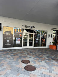 Escape-room storefront in a covered shopping plaza with glass double doors and colorful game posters, tiled pedestrian plaza with two round manhole covers and an orange planter.