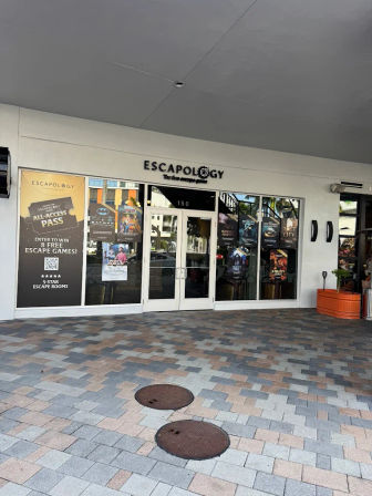 Escape-room storefront in a covered shopping plaza with glass double doors and colorful game posters, tiled pedestrian plaza with two round manhole covers and an orange planter.