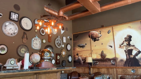 Steampunk-style café interior with wall of vintage clocks, exposed copper pipes, Edison bulbs, globe and diving helmet on the bar, and a large airship mural