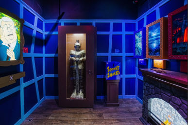 Blue-lit themed escape room featuring a knight in a glass case, a 'Scooby Snacks' dispenser, framed spooky paintings and a faux stone fireplace.