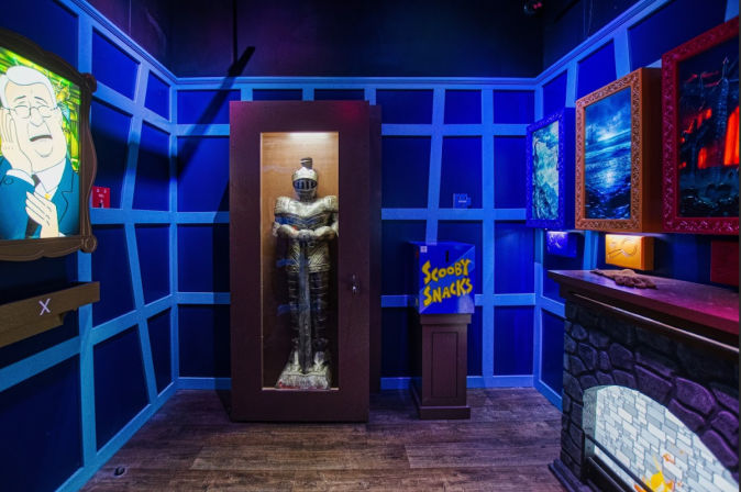 Blue-lit themed escape room featuring a knight in a glass case, a 'Scooby Snacks' dispenser, framed spooky paintings and a faux stone fireplace.