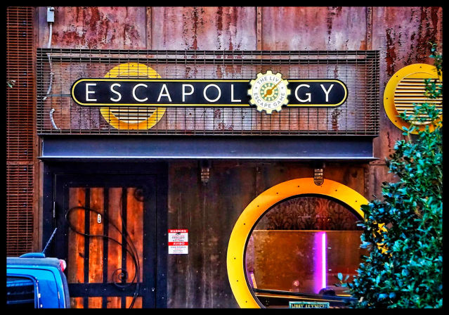 Urban escape-room storefront with industrial metal sign, rusted facade, round yellow-framed porthole window glowing purple light, wooden door with decorative ironwork, nearby greenery and a parked blue van