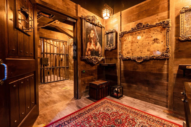 Warm, wood-paneled adventure room with ornate gold frames and vintage map, a portrait of a woman, a barred iron cell visible through the doorway, a wooden chest and red Persian-style rug â atmospheric indoor escape-room setting.