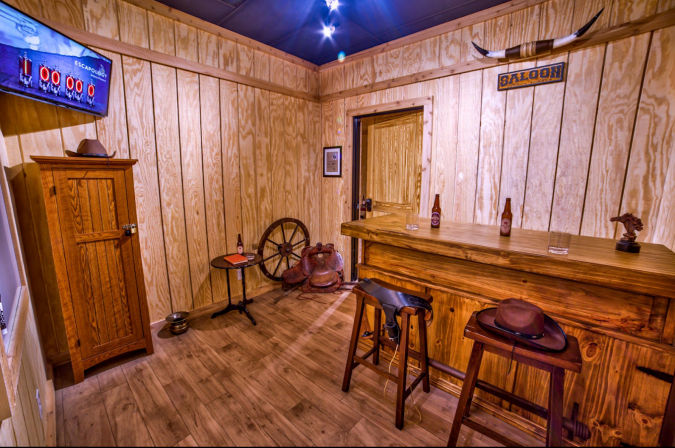 Cowboy-themed rustic saloon room with wooden paneled walls, long wooden bar and stools with beer bottles, cowboy hats, saddle, wagon wheel and mounted longhorn sign.