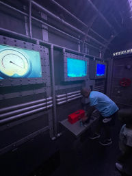 Child interacting with controls in a dimly lit submarine-style interactive museum exhibit, riveted metal panels, pressure gauge and blue porthole screens, red toolbox on the console