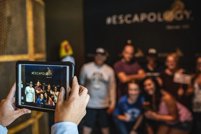 Hands holding a tablet photographing a smiling group posing in front of an escape-room backdrop inside an indoor entertainment venue.
