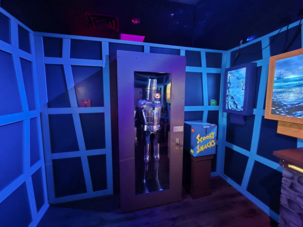 Blue-lit themed attraction room with a suit of armor in a glass display cabinet, a 'Scooby Snacks' snack box, grid-pattern walls and framed seascape artwork.