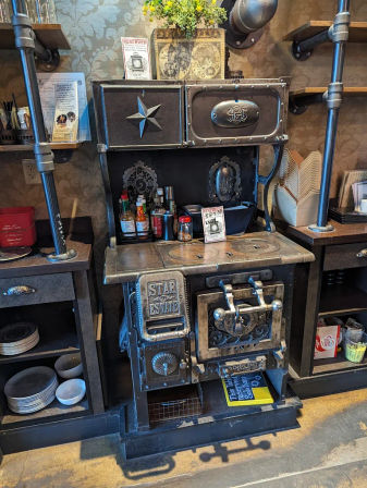 Antique cast-iron stove repurposed as a decorative service station in a cozy café interior — ornate metal doors and star emblem, condiment bottles, napkin holder, potted plant on top, and industrial pipe shelving with stacked plates nearby.