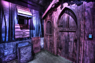 Purple-lit escape room or haunted attraction interior featuring a gothic wooden church-style door with arched window and keypad, forest mural backdrop, wooden drop box and stone-textured lower wall.