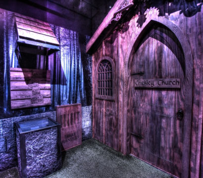 Purple-lit escape room or haunted attraction interior featuring a gothic wooden church-style door with arched window and keypad, forest mural backdrop, wooden drop box and stone-textured lower wall.
