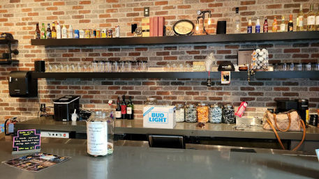 Cozy brick-walled bar interior with stainless-steel counter, floating shelves stacked with bottles and cans, rows of glassware, jars of snacks, a coffee station, tip jar and a purse on the bar.