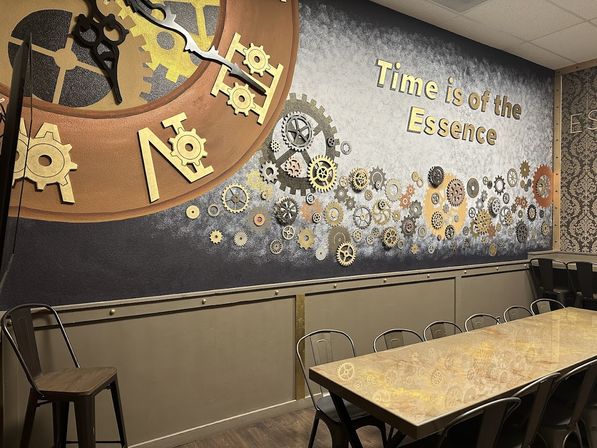 Steampunk wall mural with oversized clock, scattered brass gears and the phrase "Time is of the Essence" above a long communal table and metal chairs in a cafe-style dining area.