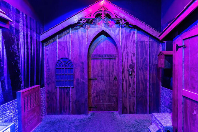 Indoor themed wooden chapel facade with arched door, decorative gable and small window, keypad and faux grass floor, bathed in vivid purple and blue LED lighting.