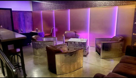 Industrial-chic lounge with four riveted metal-trimmed leather armchairs around a metal trunk coffee table, polished concrete floor, purple LED backlit wall panels and nearby bar seating