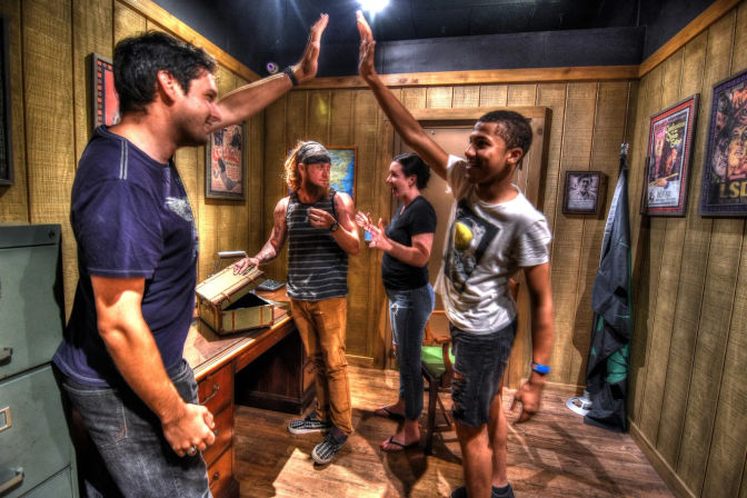 Four adults celebrate a victory in a wood-paneled escape room — two men high-five while a bearded man and a woman clap beside a desk with an open vintage chest and movie posters on the walls.