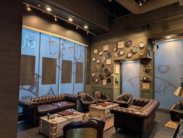 Cozy industrial steampunk lounge with tufted brown leather sofas and armchairs around vintage trunk coffee tables, a wall of assorted clocks, and large blue gear-patterned window shades.