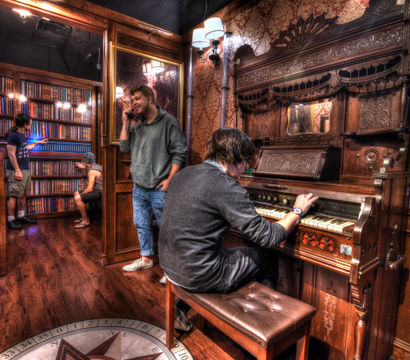 Cozy Victorian-style parlor with a person playing a carved antique pump organ, wood-paneled walls and patterned wallpaper, a book-lined alcove with others browsing, warm wall sconces and hardwood floors.