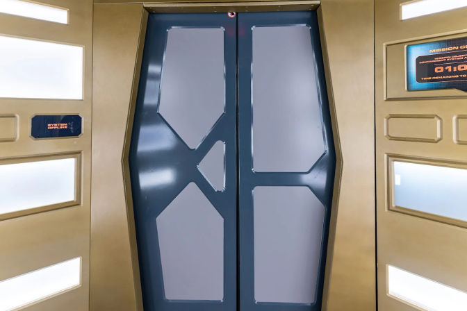 Sleek sci‑fi spaceship-style double sliding doors with dark blue geometric panels set in a gold-toned corridor wall, lit control displays and recessed white light panels