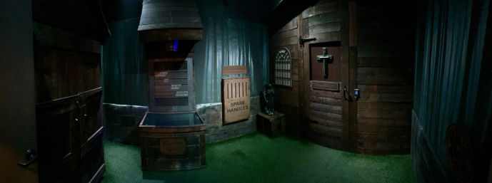 Eerie cabin-style escape room interior with wood-paneled walls, wishing well prop, crate labeled 'SPARE HANDLES', small statue, and locked door with cross and keypad.