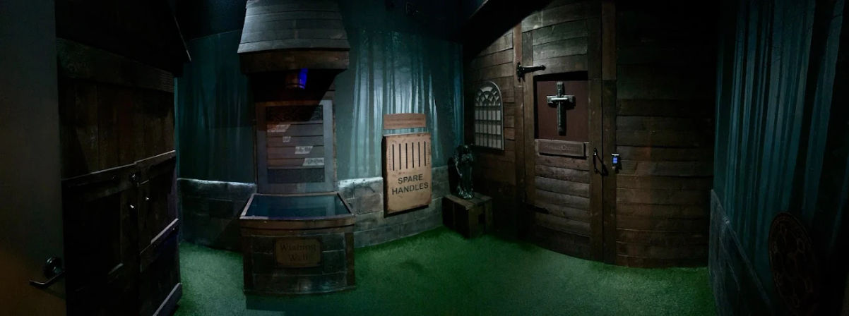 Eerie cabin-style escape room interior with wood-paneled walls, wishing well prop, crate labeled 'SPARE HANDLES', small statue, and locked door with cross and keypad.