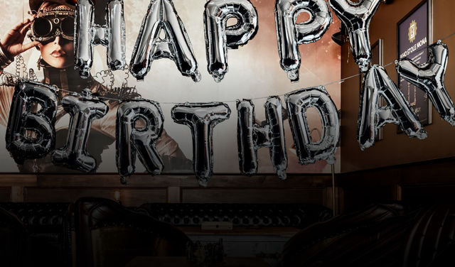 Silver foil 'HAPPY BIRTHDAY' balloons strung across a dim indoor lounge with tufted leather sofas and a large steampunk-style mural on the wall — festive bar/restaurant setting.