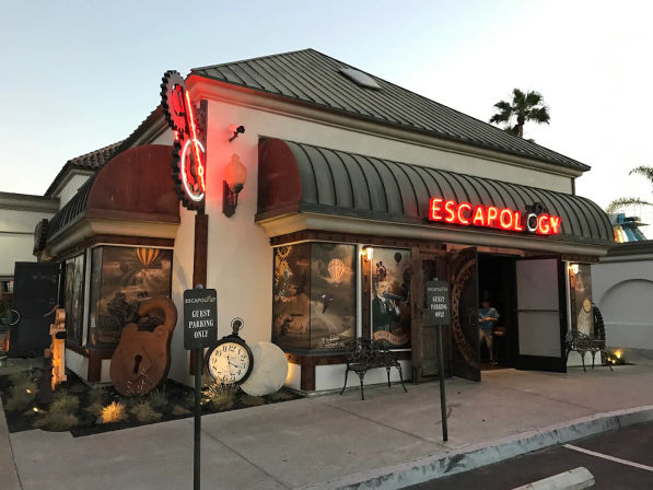Themed escape room storefront at dusk with a glowing red neon sign, curved metal awning, steampunk murals, oversized padlock and clock props, benches and palm trees in the background.
