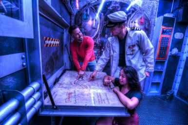 Three people in a submarine-themed escape room: a captain in a white jacket and hat points to a nautical map on a metal table while two teammates study it under moody blue lighting — immersive indoor adventure.