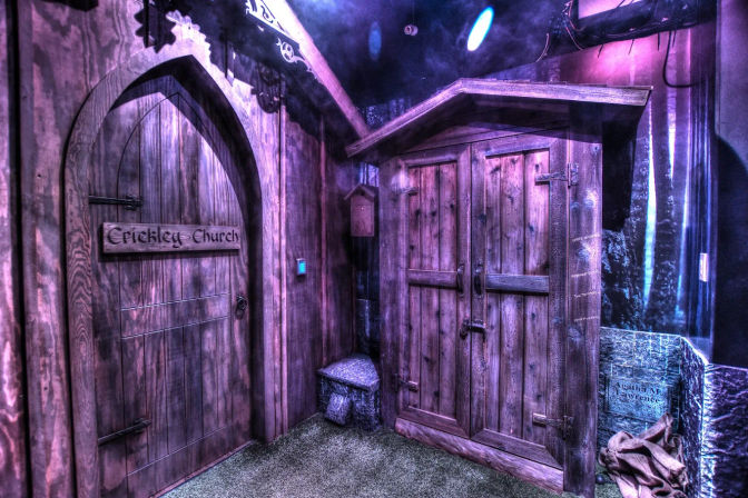 Atmospheric indoor set with an arched wooden church-style door and adjacent double-door wooden cabinet, bathed in purple-blue theatrical lighting over a grassy floor with a stone bench and rustic props.