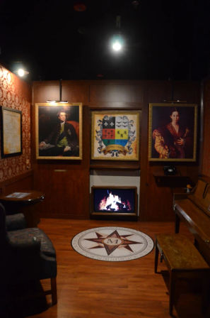Historic-style parlor interior with wood-paneled walls, two oil portraits and a heraldic coat of arms above a glowing fireplace, compass medallion on the hardwood floor, piano bench and upholstered armchair.