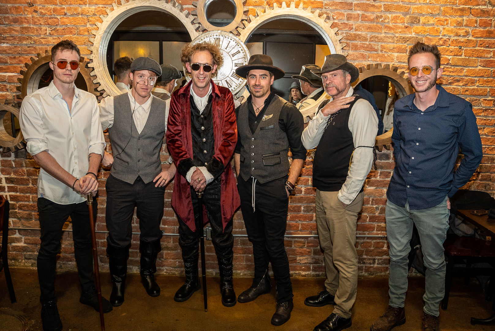 Six men in steampunk and vintage-inspired outfits — hats, canes and round sunglasses — pose for a group portrait against an exposed brick wall with oversized industrial gears and a clock.