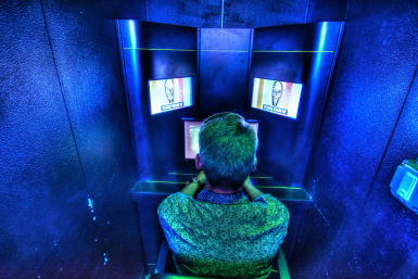 Person seen from behind seated in a small neon-blue kiosk, leaning toward three glowing screens in a cyber-arcade style booth.